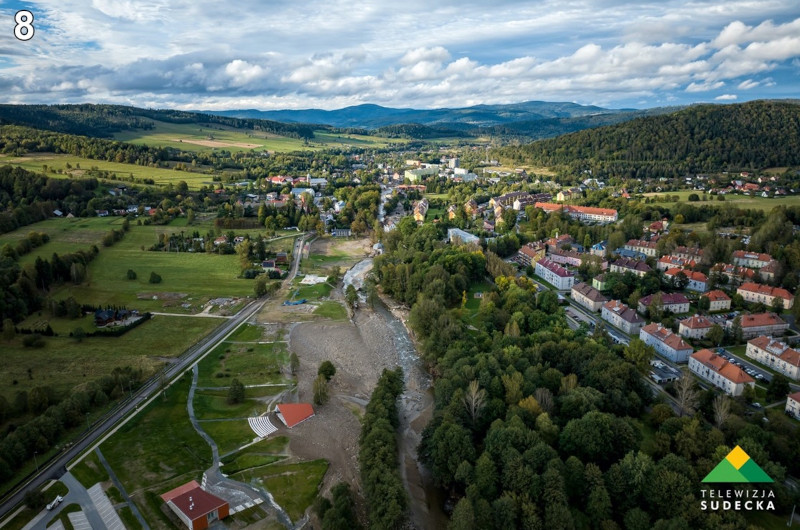 Widok z lotu ptaka na malowniczy krajobraz. Widać na nim rozległe tereny zielone, pola, lasy oraz zabudowania miejskie. W dolnej części zdjęcia znajdują się budynki mieszkalne, a w tle rozciągają się wzgórza i góry. Na niebie widoczne są chmury. W lewym dolnym rogu zdjęcia znajduje się logo "Telewizja Sudecka".