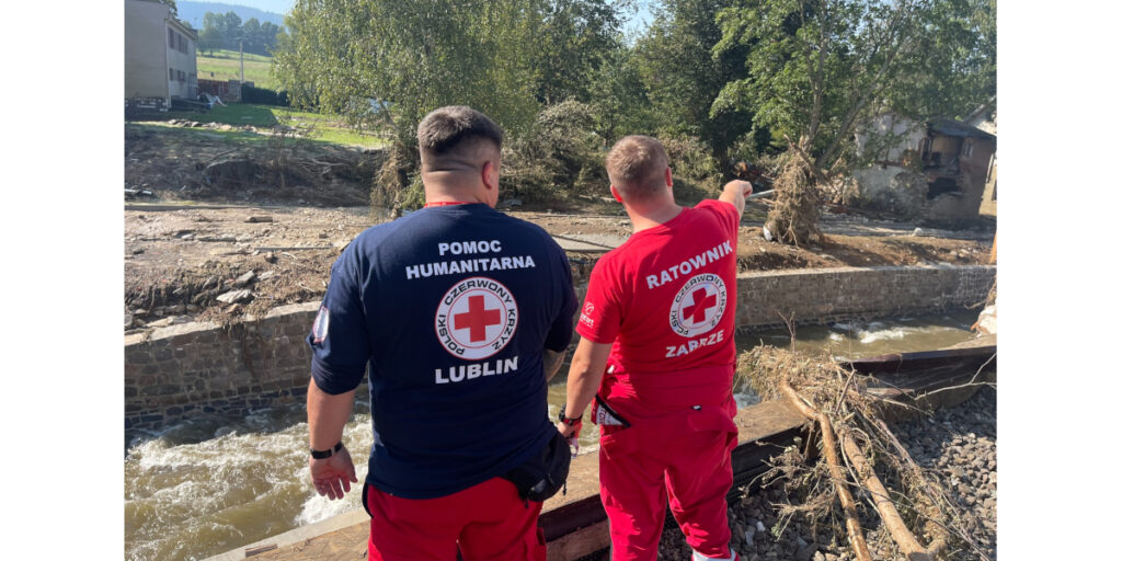 Na pierwszym planie dwóch mężczyzn stoi tyłem. Mężczyzna z prawej strony ma jasne włosy, czerwoną koszulkę i czerwone spodnie. Na koszulce logo PCK. Nad logiem napis 