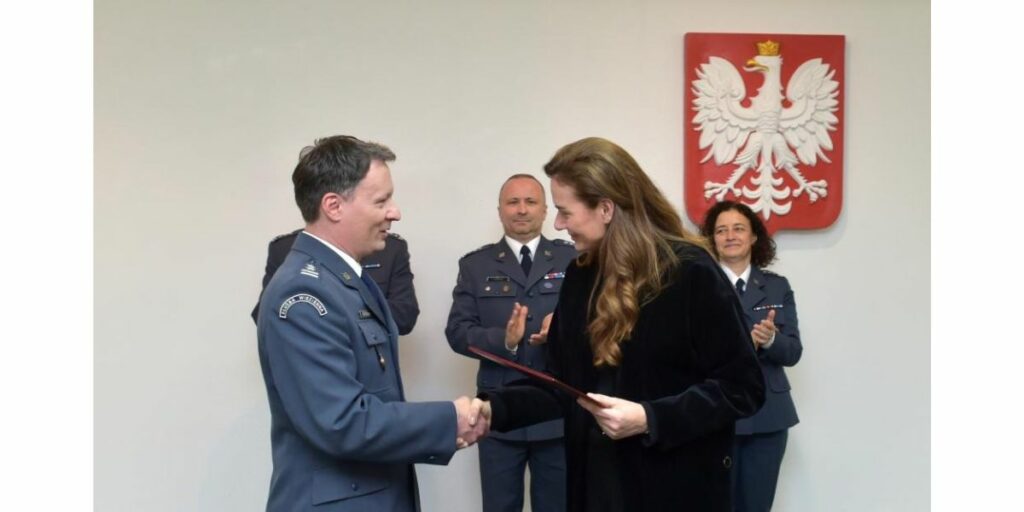 Uroczystość. Pięć osób, cztery w mundurach służby więziennej. Piąta to wiceminister sprawiedliwości Maria Ejchart ubrana na czarno, stojąca bokiem. Podaje rękę mężczyźnie w mundurze. Ten mężczyzna to podpułkownik Cezary Mecwaldowski. Kobieta wręcza mu dokument i gratuluje awansu na zastępcę dyrektora generalnego Służby Więziennej. W tle na ścianie wisi godło Polski. Pozostałe trzy osoby w mundurach stoją z tyłu. Klaszczą.