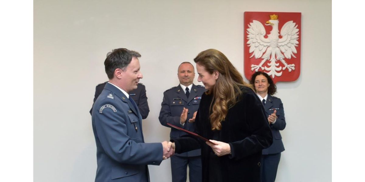 Uroczystość. Pięć osób, cztery w mundurach służby więziennej. Piąta to wiceminister sprawiedliwości Maria Ejchart ubrana na czarno, stojąca bokiem. Podaje rękę mężczyźnie w mundurze. Ten mężczyzna to podpułkownik Cezary Mecwaldowski. Kobieta wręcza mu dokument i gratuluje awansu na zastępcę dyrektora generalnego Służby Więziennej. W tle na ścianie wisi godło Polski. Pozostałe trzy osoby w mundurach stoją z tyłu. Klaszczą.