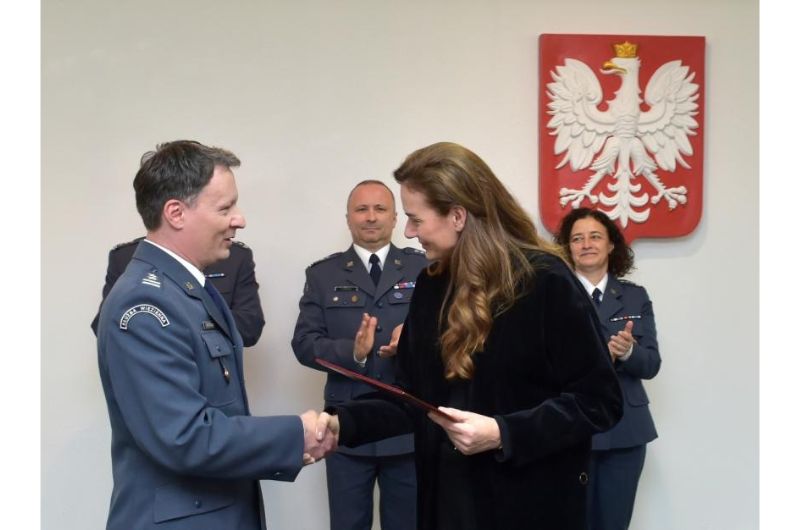 Uroczystość. Pięć osób, cztery w mundurach wojskowych. Piąta to wiceminister sprawiedliwości Maria Ejchart ubrana na czarno, stojąca bokiem. Podaje rękę mężczyźnie w mundurze. Ten mężczyzna to podpułkownik Cezary Mecwaldowski. Kobieta wręcza mu dokument i gratuluje awansu na zastępcę dyrektora generalnego Służby Więziennej. W tle na ścianie wisi godło Polski. Pozostałe trzy osoby w mundurach stoją z tyłu. Klaszczą.