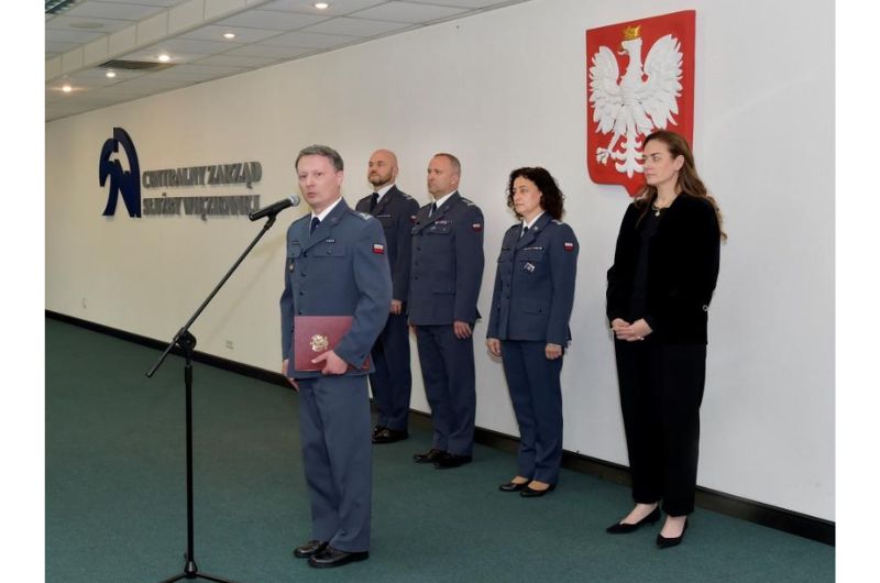 Awans ppłk. Cezarego Mecwaldowskiego na zastępcę dyrektora generalnego Służby Więziennej. Pięć osób. Cztery w mundurach służby więziennej. Piąta to wiceminister sprawiedliwości Maria Ejchart ubrana na czarno, Pani wiceminister i trzy osoby w mundurach stoją przy ścianie. Tyłem do nich i bokiem do aparatu przy mikrofonie stoi podpułkownik Cezary Mecwaldowski. W lewej ręce trzyma bordową teczkę z godłem Polski. W tle na ścianie widnieje logo Centralnego zarządu Służby Więziennej. Nad głowami osób stojących przy ścianie wisi godło Polski.