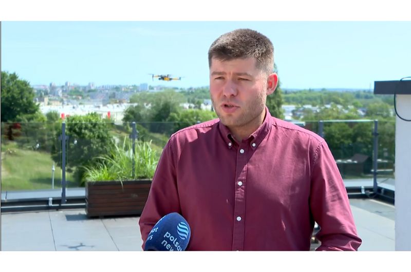Kadr z programu Polsat News. Sergiusz Parszowski w bordowej koszuli stojący na zewnątrz mówi do mikrofonu, który trzyma niewidoczna druga osoba. W tle krajobraz miejski: dachy budynków i drzewa. Na niebie lecący dron.