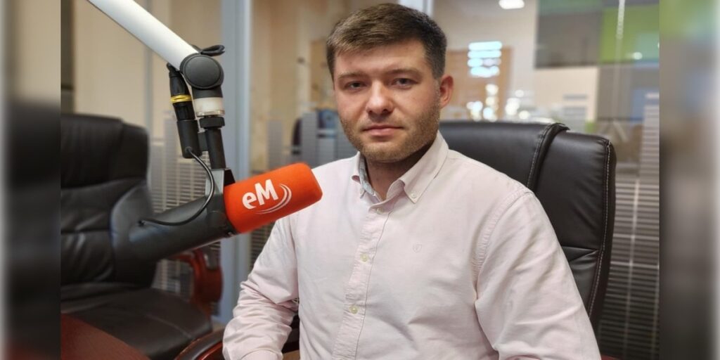 Mężczyzna siedzi na skórzanym fotelu w studiu radiowym, ubrany w jasną koszulę. Przed nim stoi mikrofon z pomarańczową osłonką i logo 