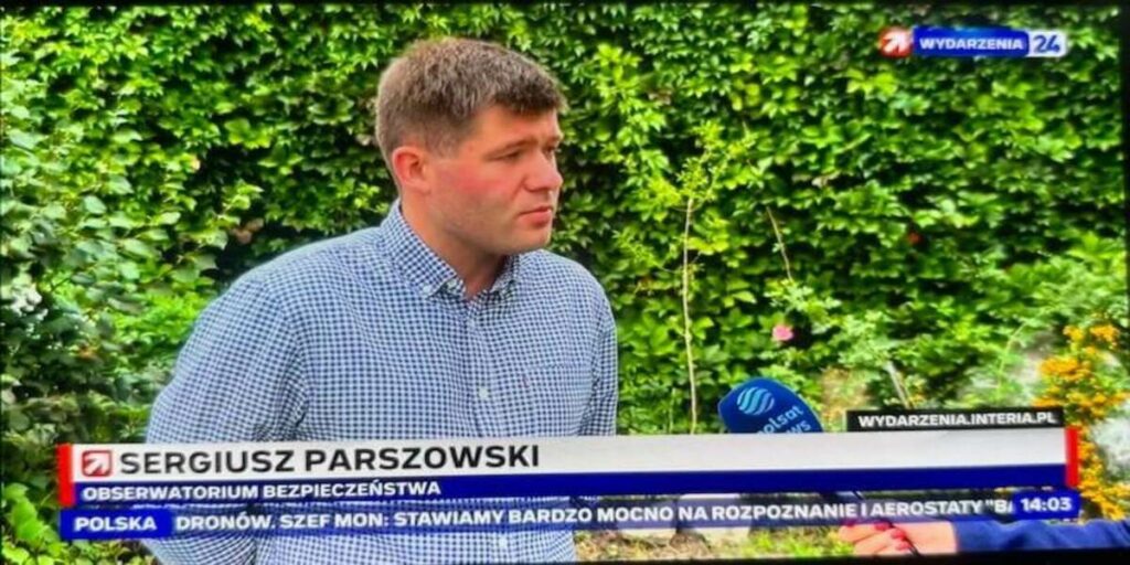 Mężczyzna ubrany w koszulę w granatowo-białą kratę stoi na zewnątrz, a w tle widać gęstą, zieloną ścianę z liści. Udziela wywiadu, a na ekranie widnieje podpis: "Sergiusz Parszowski, Obserwatorium Bezpieczeństwa". Obok niego znajduje się mikrofon Polsat News.