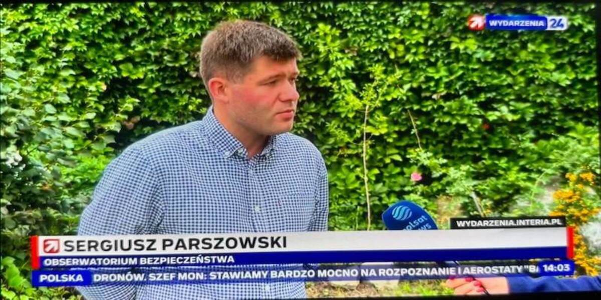 Mężczyzna ubrany w koszulę w granatowo-białą kratę stoi na zewnątrz, a w tle widać gęstą, zieloną ścianę z liści. Udziela wywiadu, a na ekranie widnieje podpis: "Sergiusz Parszowski, Obserwatorium Bezpieczeństwa". Obok niego znajduje się mikrofon Polsat News.