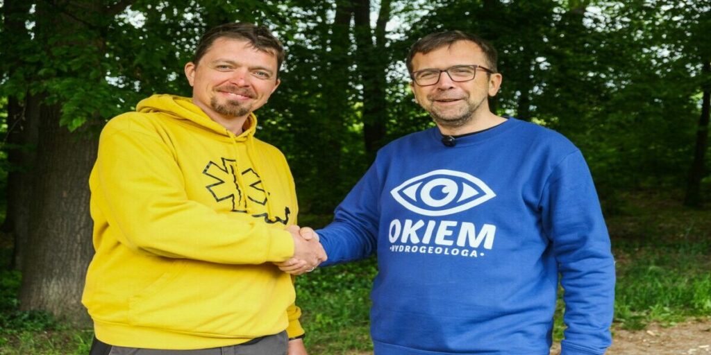 Dwóch mężczyzn, jeden w żółtej bluzie z kapturem, drugi w niebieskiej bluzie z napisem 'OKIEM HYDROGEOLOGA' i symbolem oka, uściska sobie dłonie w plenerze, na tle drzew