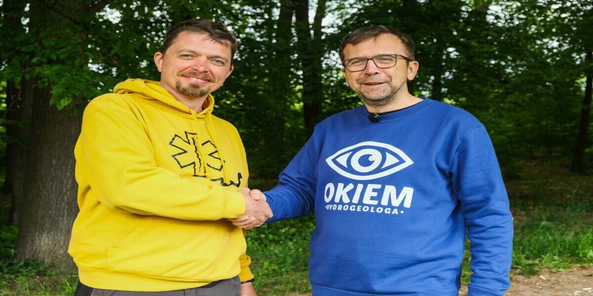 Dwóch mężczyzn, jeden w żółtej bluzie z kapturem, drugi w niebieskiej bluzie z napisem 'OKIEM HYDROGEOLOGA' i symbolem oka, uściska sobie dłonie w plenerze, na tle drzew