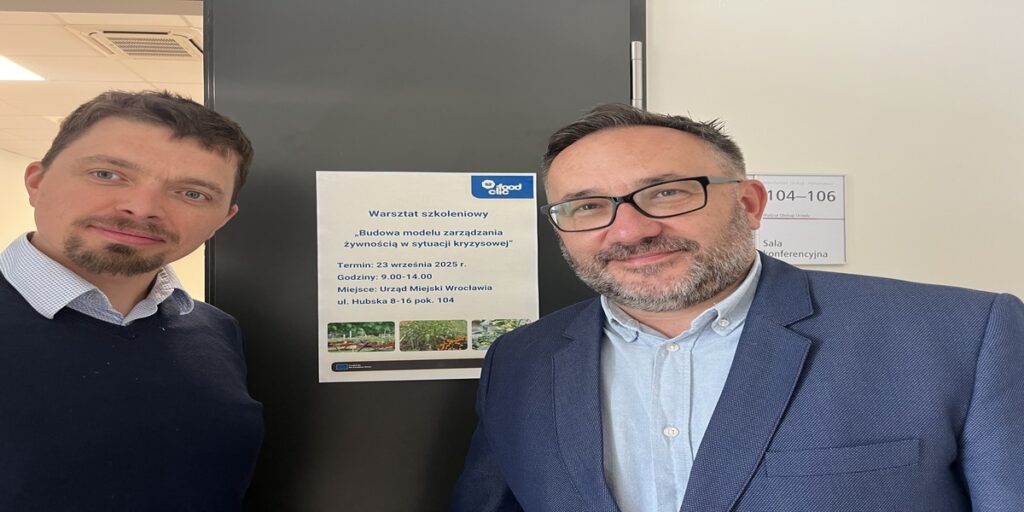 Dwóch mężczyzn stoi obok informacji o warsztatach projektu FoodCLIC, zatytułowanych „Budowa modelu zarządzania żywnością w sytuacji kryzysowej”, które odbywają się w Urzędzie Miejskim Wrocławia.