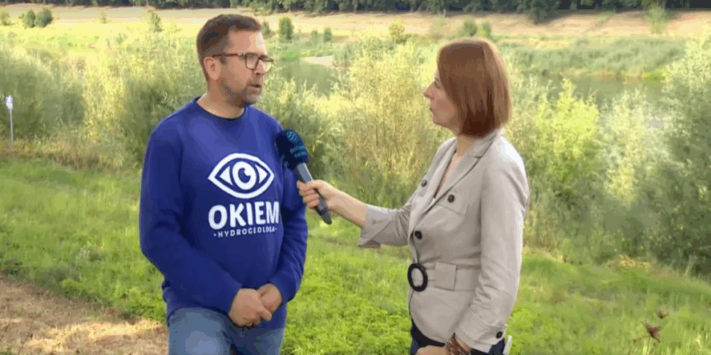 Kobieta-reporterka w jasnej kurtce, trzymająca mikrofon z logo stacji telewizyjnej, przeprowadza wywiad z mężczyzną w okularach i niebieskiej bluzie z napisem "OKIEM HYDROGEOLOGA". Rozmawiają na zewnątrz, na tle rzeki i zielonych zarośli.